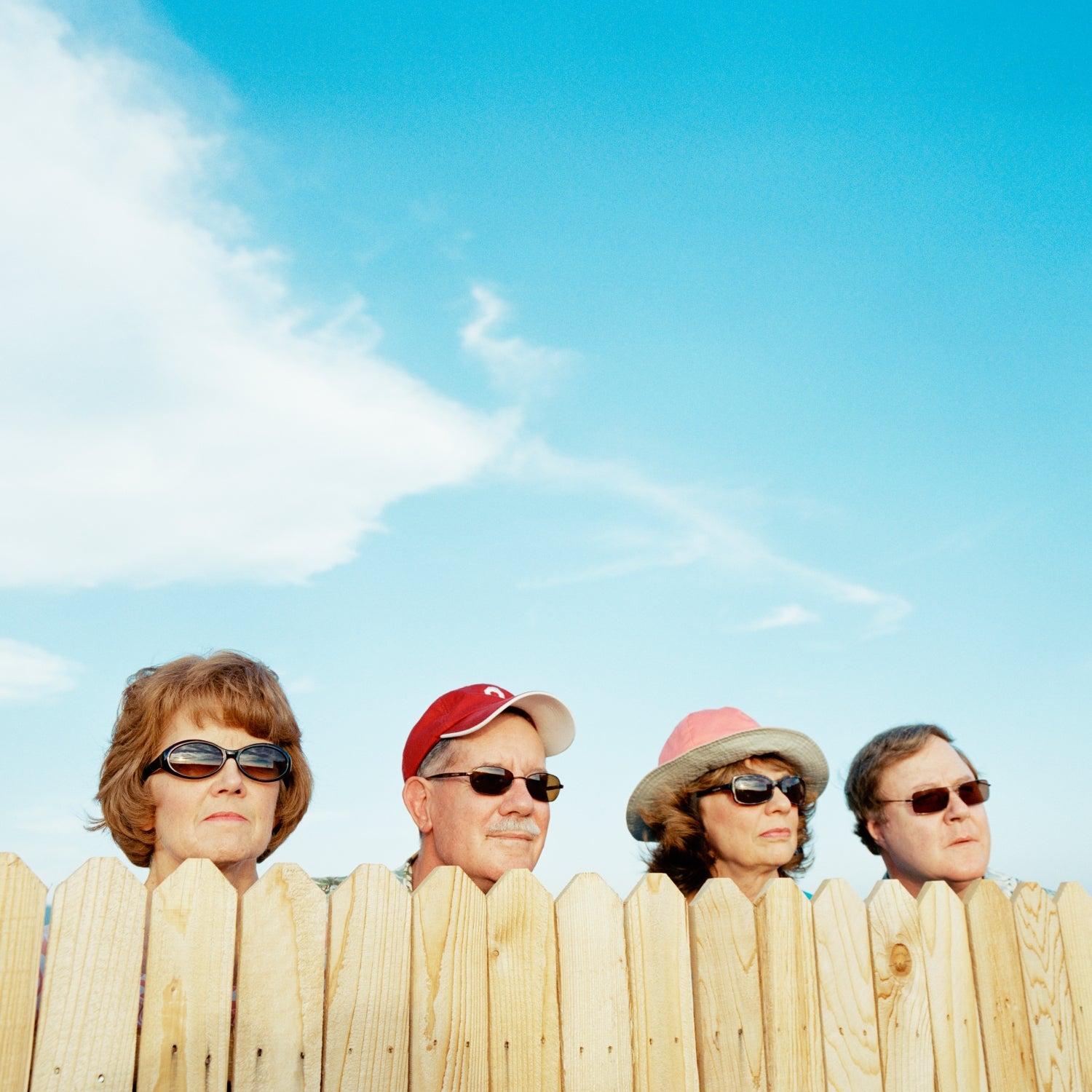 Four older neighbors wearing sunglasses peer over a wooden fence