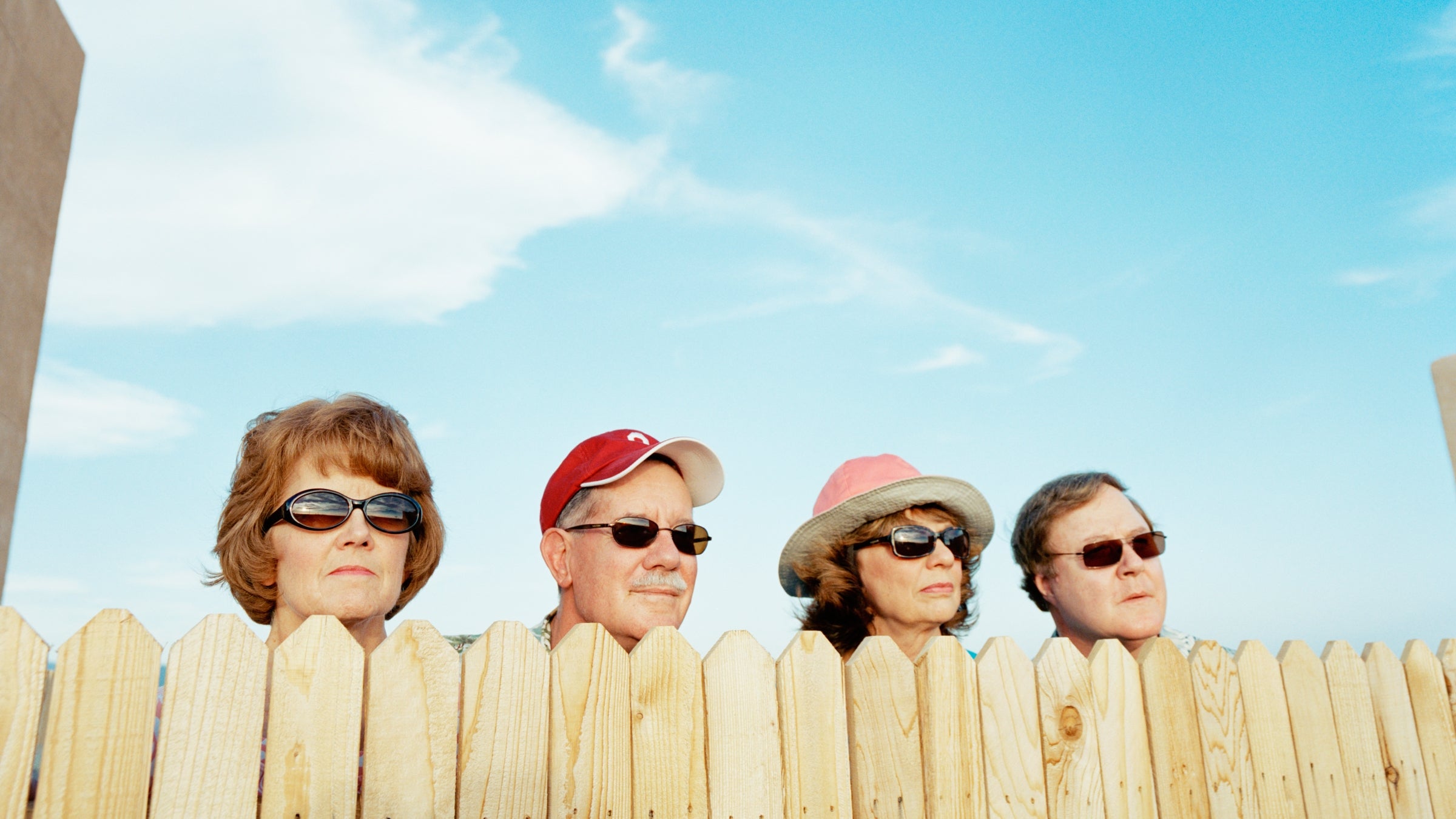 Four older neighbors wearing sunglasses peer over a wooden fence