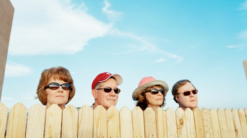 Four older neighbors wearing sunglasses peer over a wooden fence