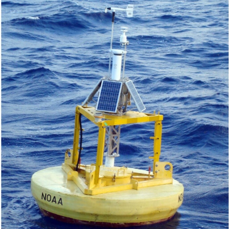 A floating yellow buoy with what appear to be solar panels atop a short metal construction floats in the ocean.