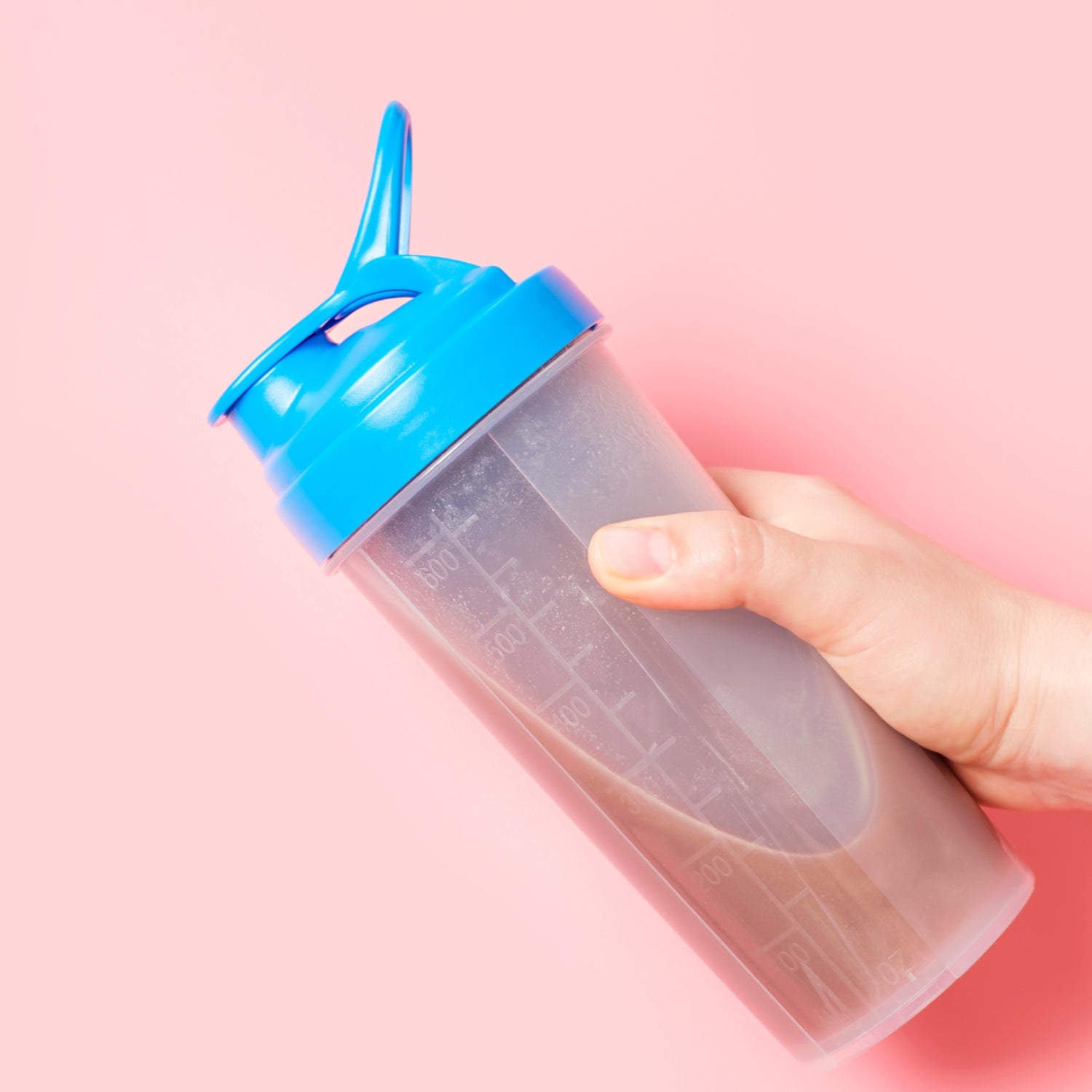 A hand holding a protein shake with a blue cap against a pale pink background