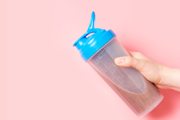 A hand holding a protein shake with a blue cap against a pale pink background
