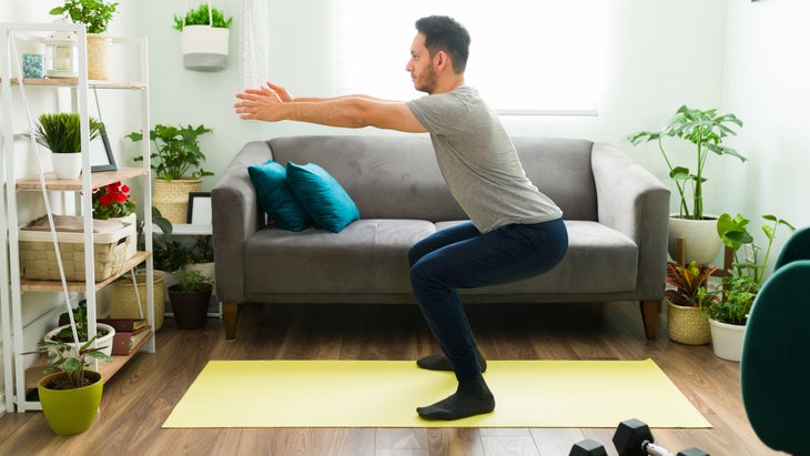 man in living room completes bodyweight workout plan by squatting with his arms straight out in front of him