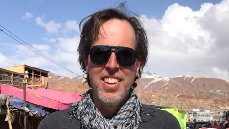 The author wearing a scarf and standing in front of a brown hillside with blue sky and clouds behind it