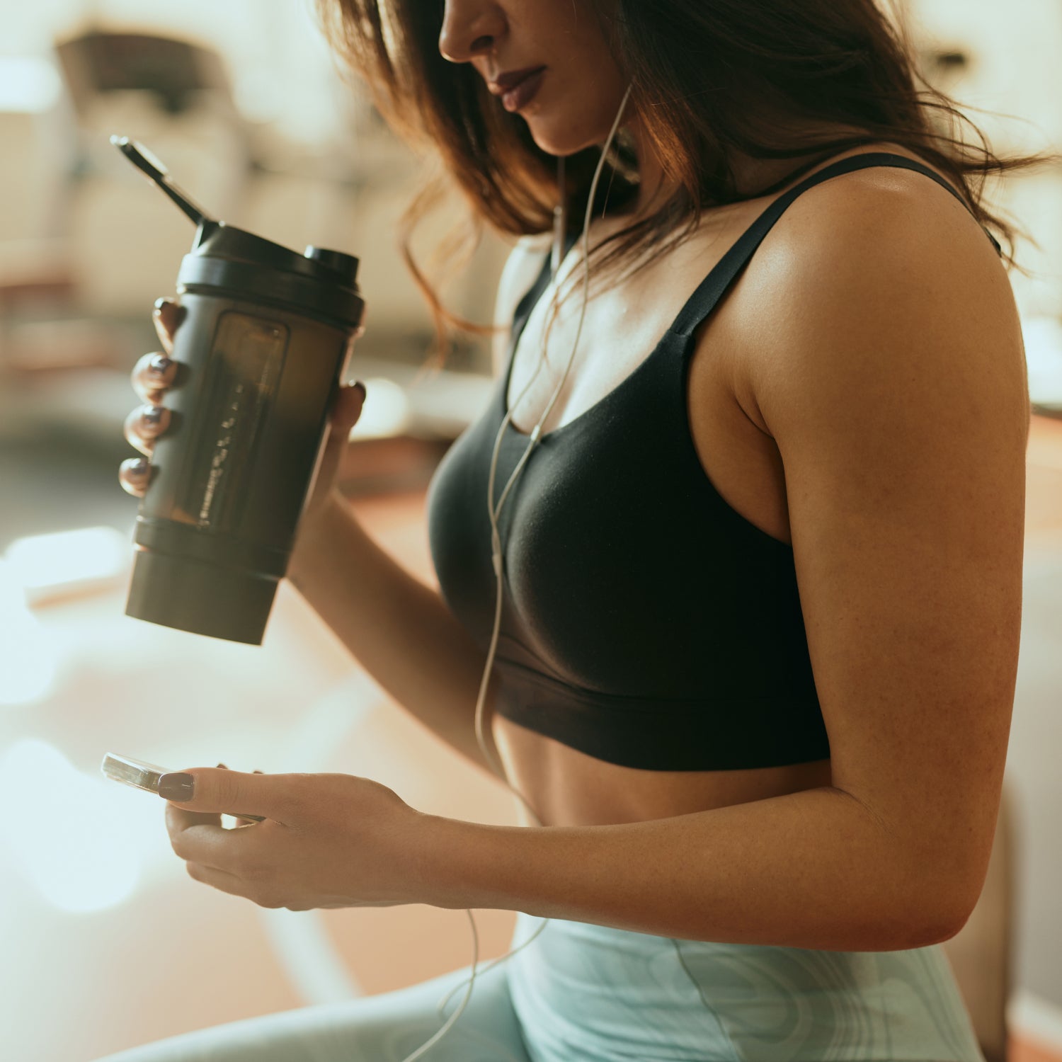 Cropped picture of a strong sportswoman sits in a gym, drinks protein drink and using a phone while drinking protein drink and taking a break.