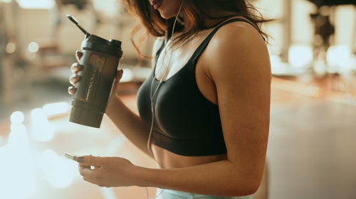 Cropped picture of a strong sportswoman sits in a gym, drinks protein drink and using a phone while drinking protein drink and taking a break.