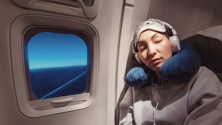 A woman wearing a neck pillow and a sleeping mask on her forehead dozes with her head leaning against the side of the inside of a plane, near a window