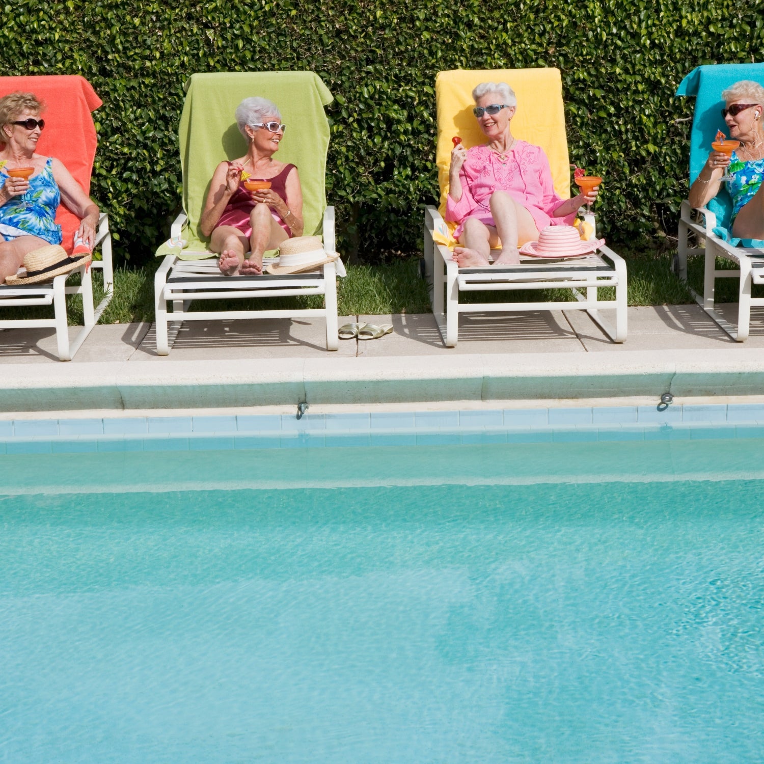 Four elderly women relax by a pool and drink cocktails