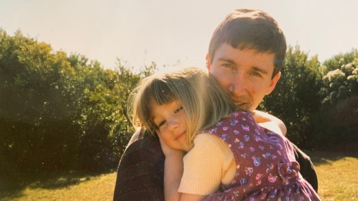 A man holds his young daughter and looks at the camera in the sunshine with bushes in the background