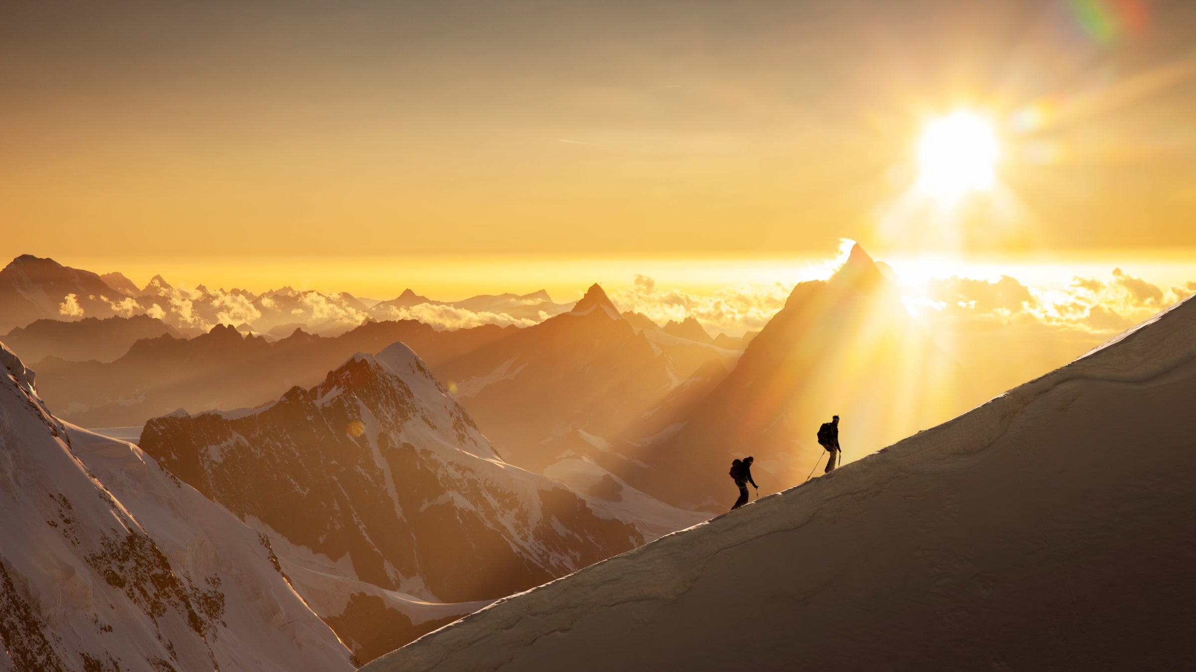 Two skiers ascending a ridgeline in the Swiss Alps at sunrise, with a jagged mountain range behind them
