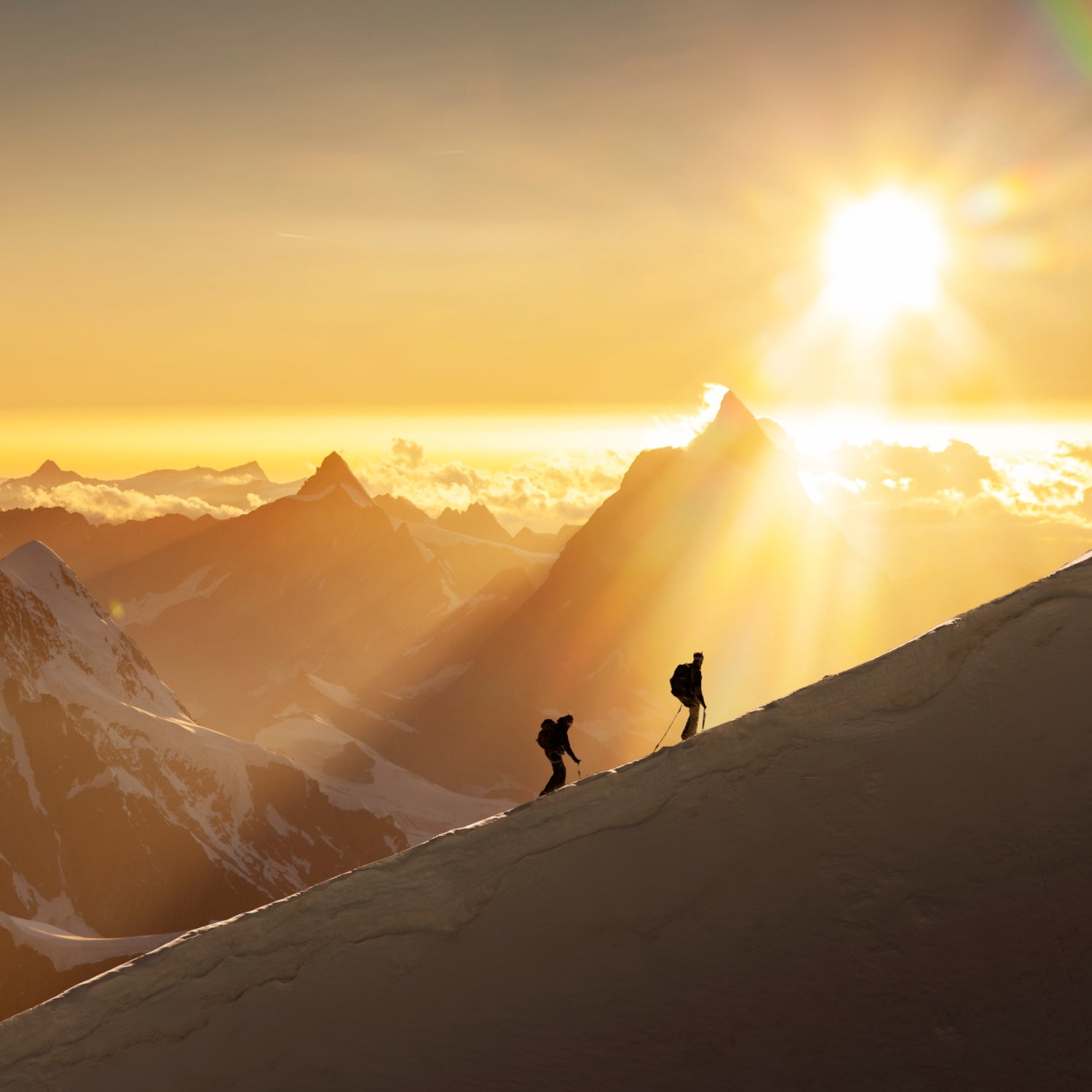 Two skiers ascending a ridgeline in the Swiss Alps at sunrise, with a jagged mountain range behind them