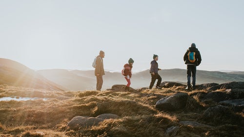A father enjoys a view with his three young children. They have a sense of achievement after scaling a mountain. The sun is low, the sky providing a space for copy.