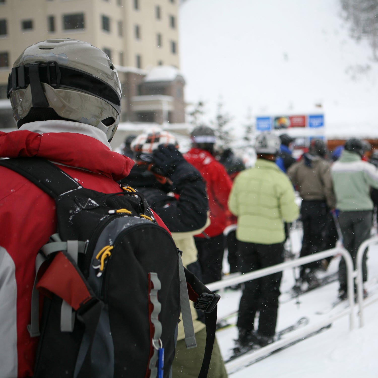 Skiers and snowboarders waiting in line for chair lift with falling snow.