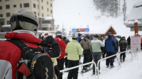 Skiers and snowboarders waiting in line for chair lift with falling snow.