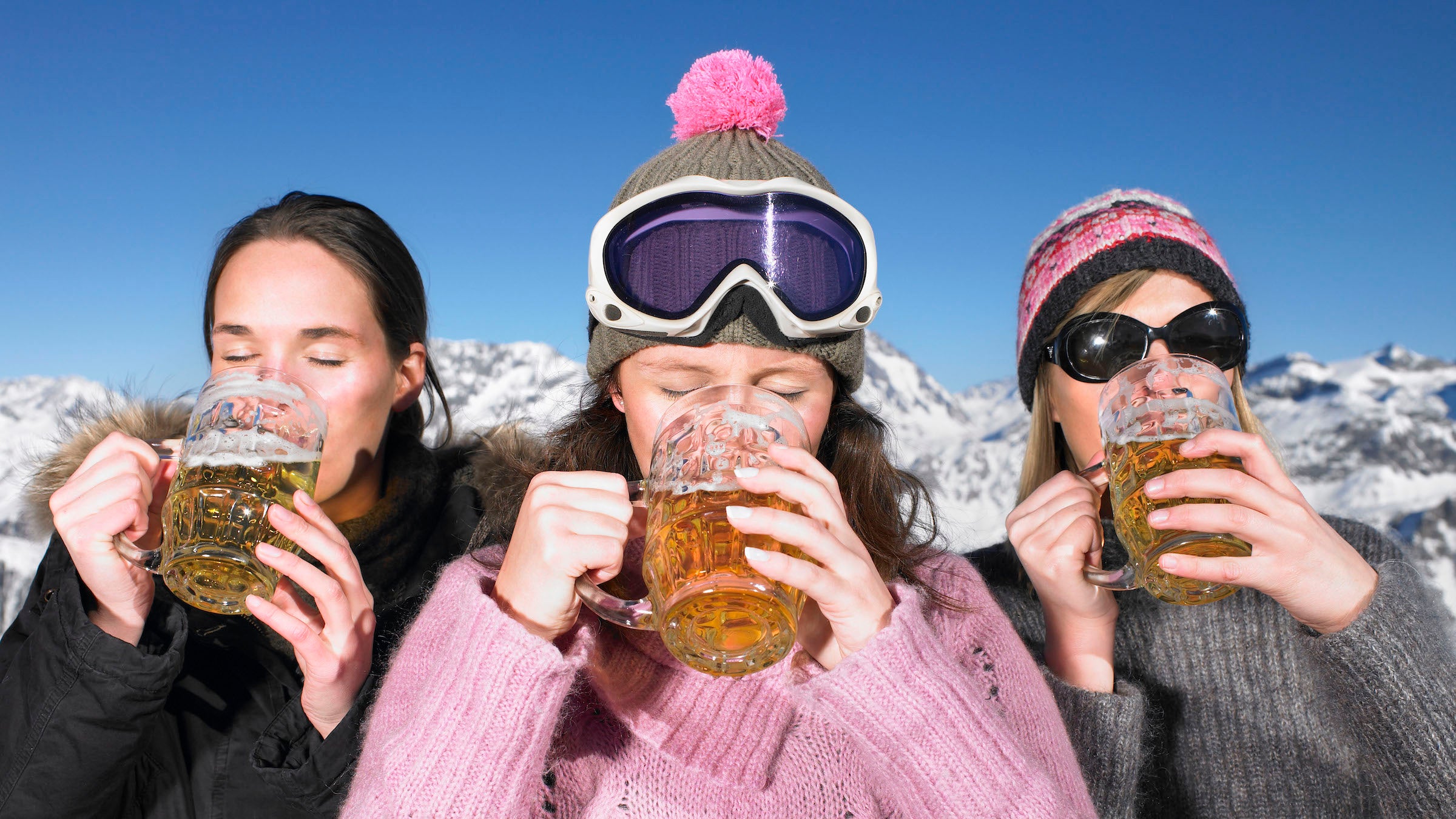 Three women in ski attire drink large steins of beer