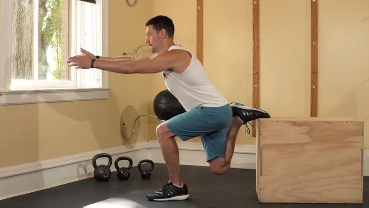 A person doing a Bulgarian split squat. Their left leg is bent at a 90-degree angle and their right leg is bent behind them, with the top of their right foot placed on a box. Their arms are outstretched in front of them.