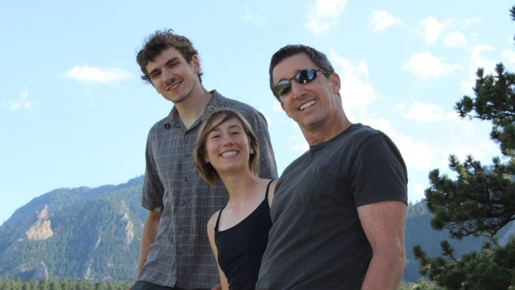 two men and a woman smile at the camera in casual clothes with a mountain in the background