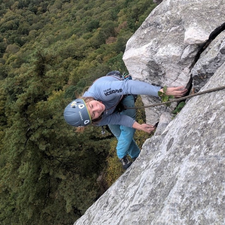 A smiling young woman in a jacket, leggings, and climbing helmet climbs a grey rock face with green trees below. 