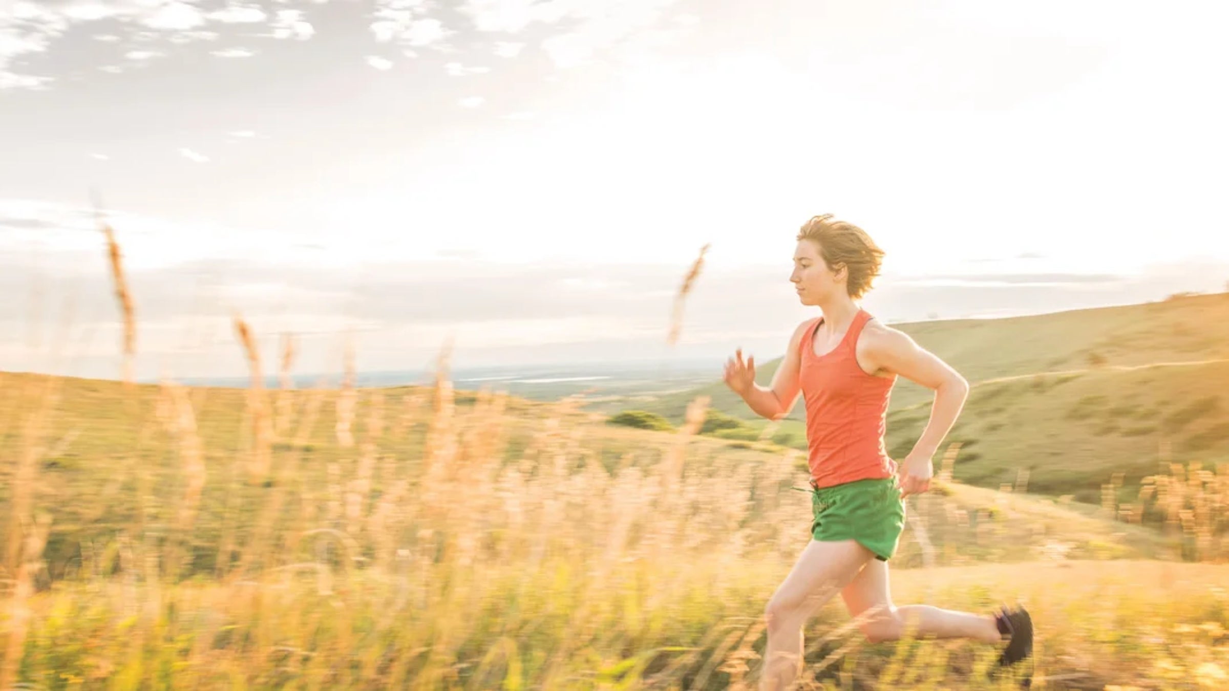 A woman runs through a golden field of grass wearing green running shorts and an orange tank top