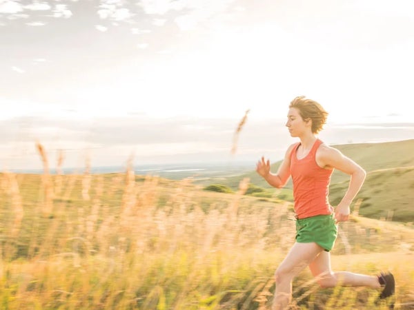 A woman runs through a golden field of grass wearing green running shorts and an orange tank top