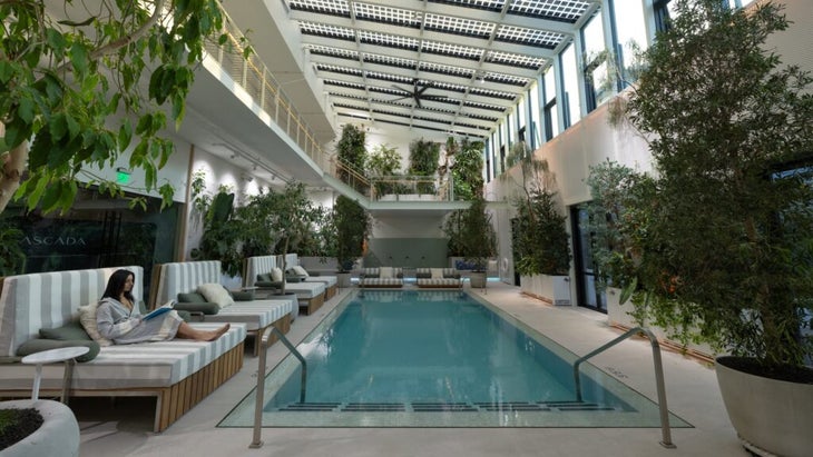 The sunlit indoor conservatory at Cascada, a new hotel in Portland, Oregon, has a rectangular-shaped pool, several trees, a windowed ceiling and chase-like chairs. 