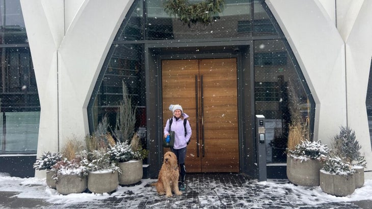 A woman dressed in cold-weather gear, posing with a big dog outside Denver's Populus hotel entrance while it snows