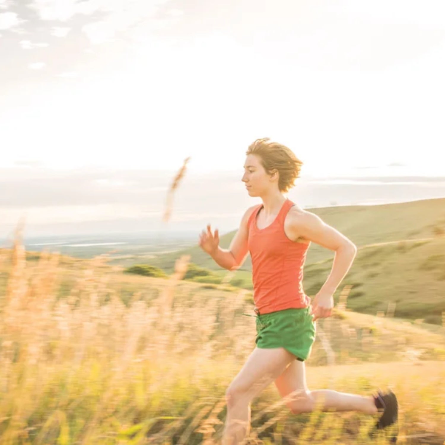 A woman runs through a golden field of grass wearing green running shorts and an orange tank top