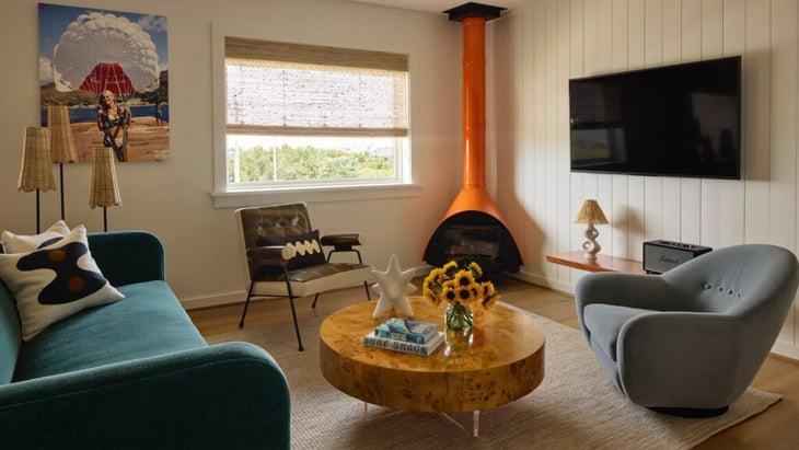 A living room of one of the rooms of Pamlico Station, a new hotel in North Carolina's Outer Banks; in the room is an orange corner woodstock, a couch, a window with a view of greenery, and a print of a girl parasailing.