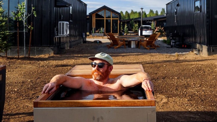 A man wearing a ball cap and sunglasses sits on a square-shaped hot tub outdoors, with some of the cabins of the Yellowstone Peaks hotel in the background