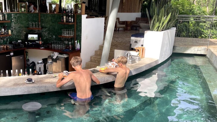 Two teenage boys sit on underwater stools at Suitree Experience’s swim-up bar, enjoying breakfast