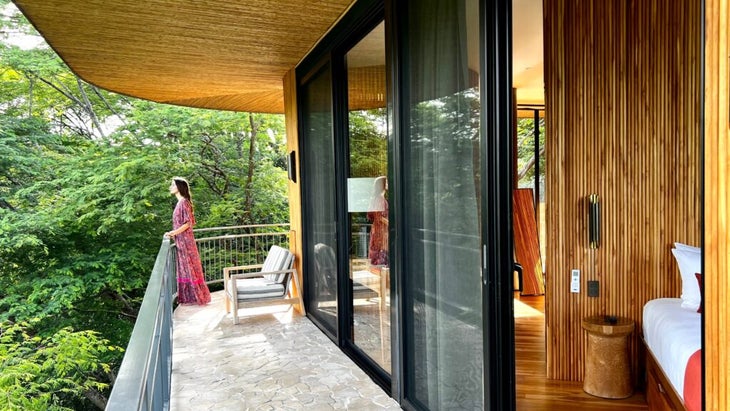 A woman stands on the porch of her Suitree room, looking at the jungle surrounds. We also see the floor-to-ceiling windows and some of the room's interior.