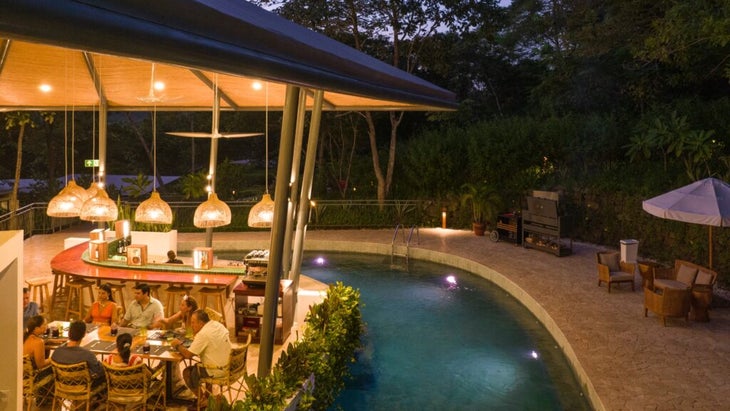 A group of six people sit at a table at Suitree’s restaurant, Taru, at dusk; you can see an adjacent pool and deck with tables and chairs