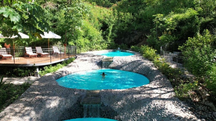 A woman swims in the middle of three connected pools, surrounded by lush foliage.