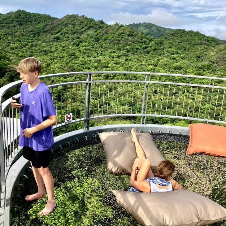 A teenage boy stands, and his mom lays back, on an outdoor deck at the Suitree Experience Hotel in Costa Rica, with views of the green jungle.