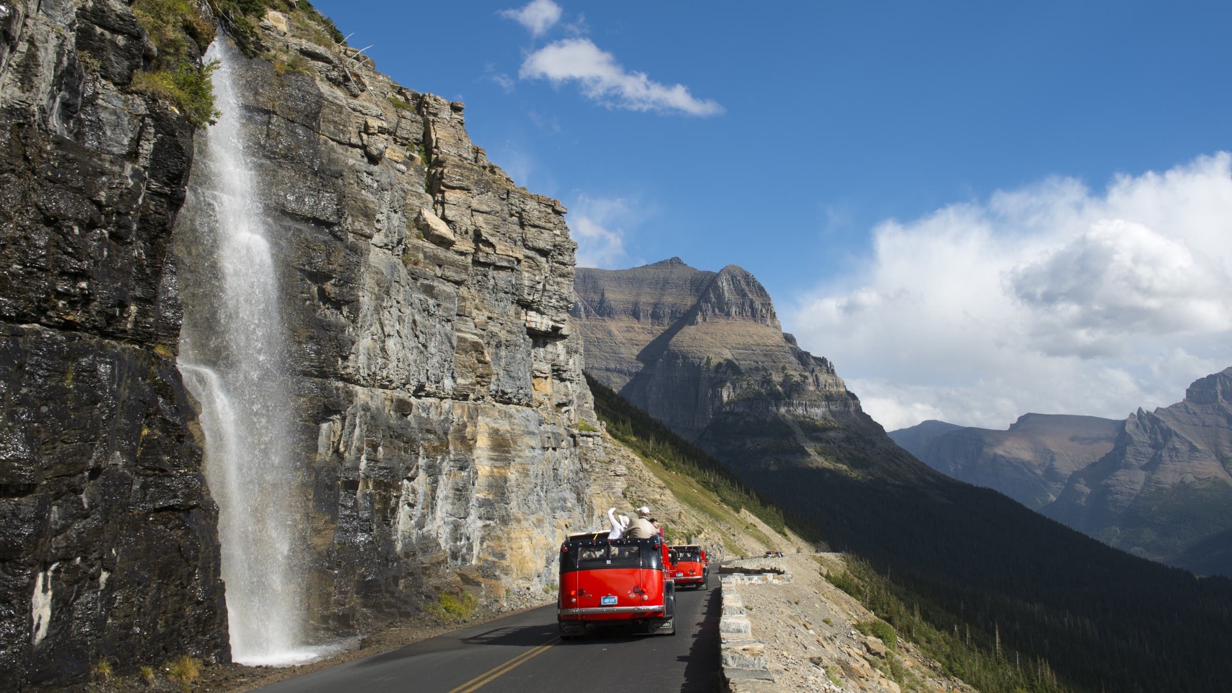 Red tour bus at waterfall coming down next to the Going-to-the-Sun Road near Logan Pass in Glacier National Park, Montana, United States.
