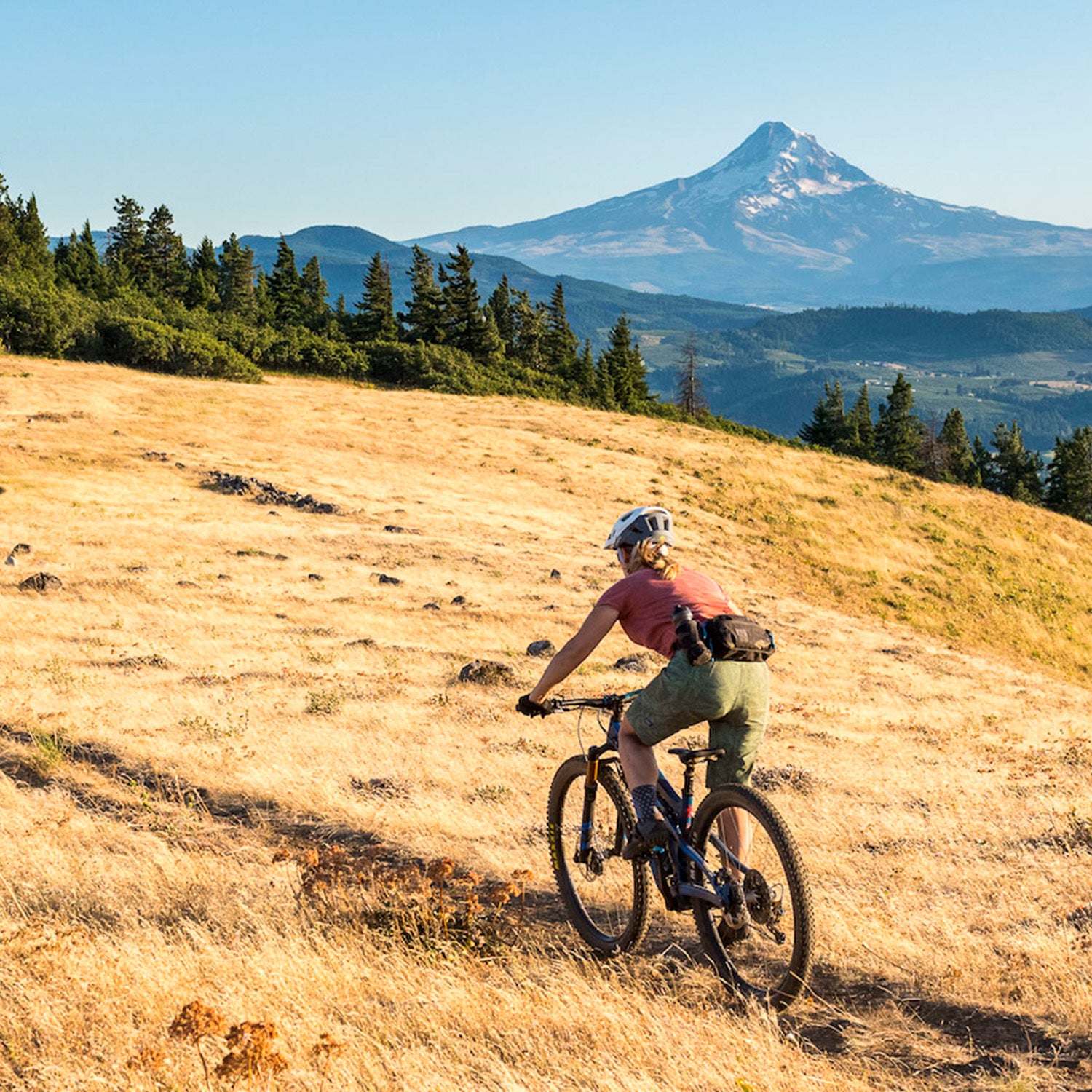 woman riding a bike across a golden grassy field with a mountain in the distance; she's wearing a hip pack