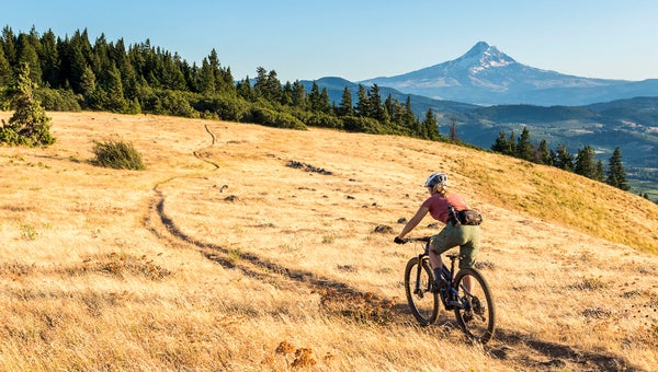 woman riding a bike across a golden grassy field with a mountain in the distance; she's wearing a hip pack