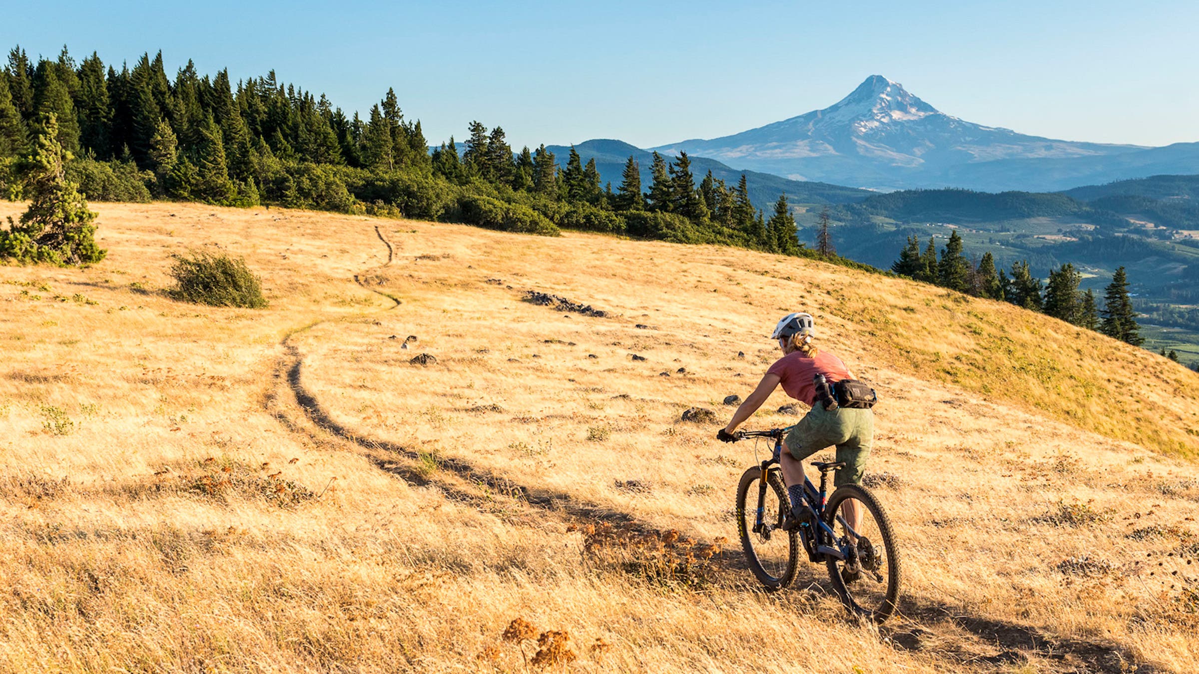Woman riding a mountain bike across a golden grassy field with a mountain in the distance; she's wearing a hip pack
