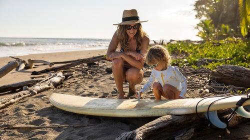 Woman crouching over toddler on surfboard on sandy beach