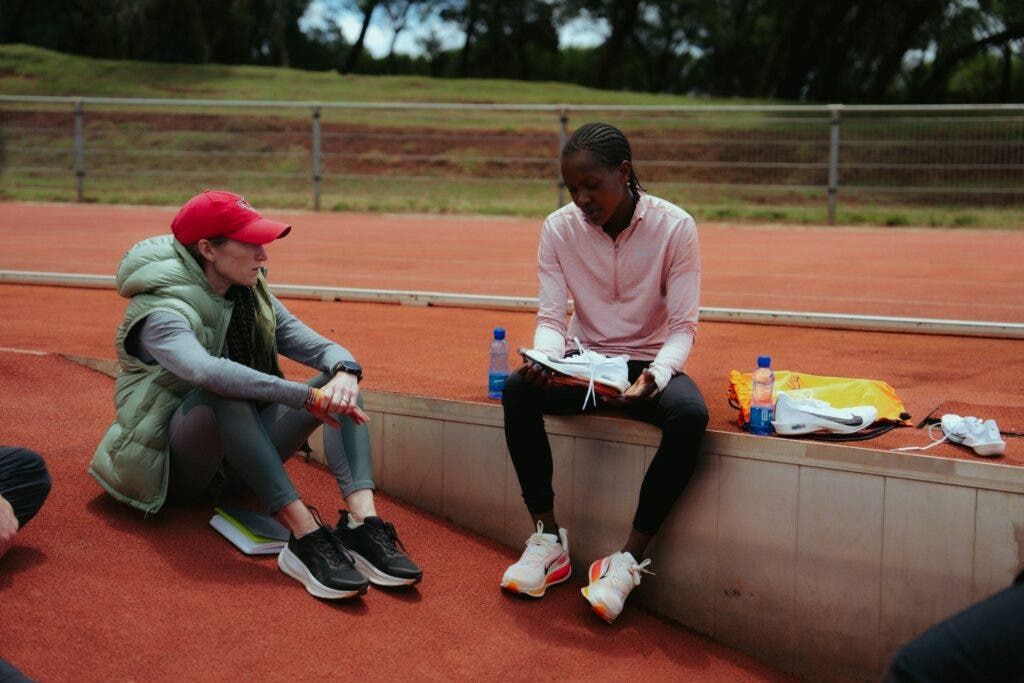 Faith Kipyegon holds a running shoe while sitting next to a racing track.