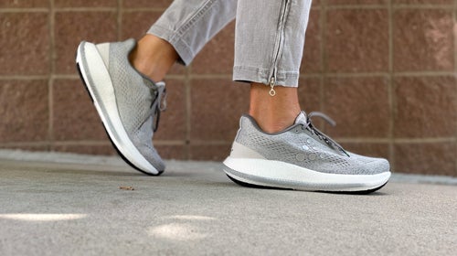 woman's feet in gray running shoes walking on pavement in front of brick wall