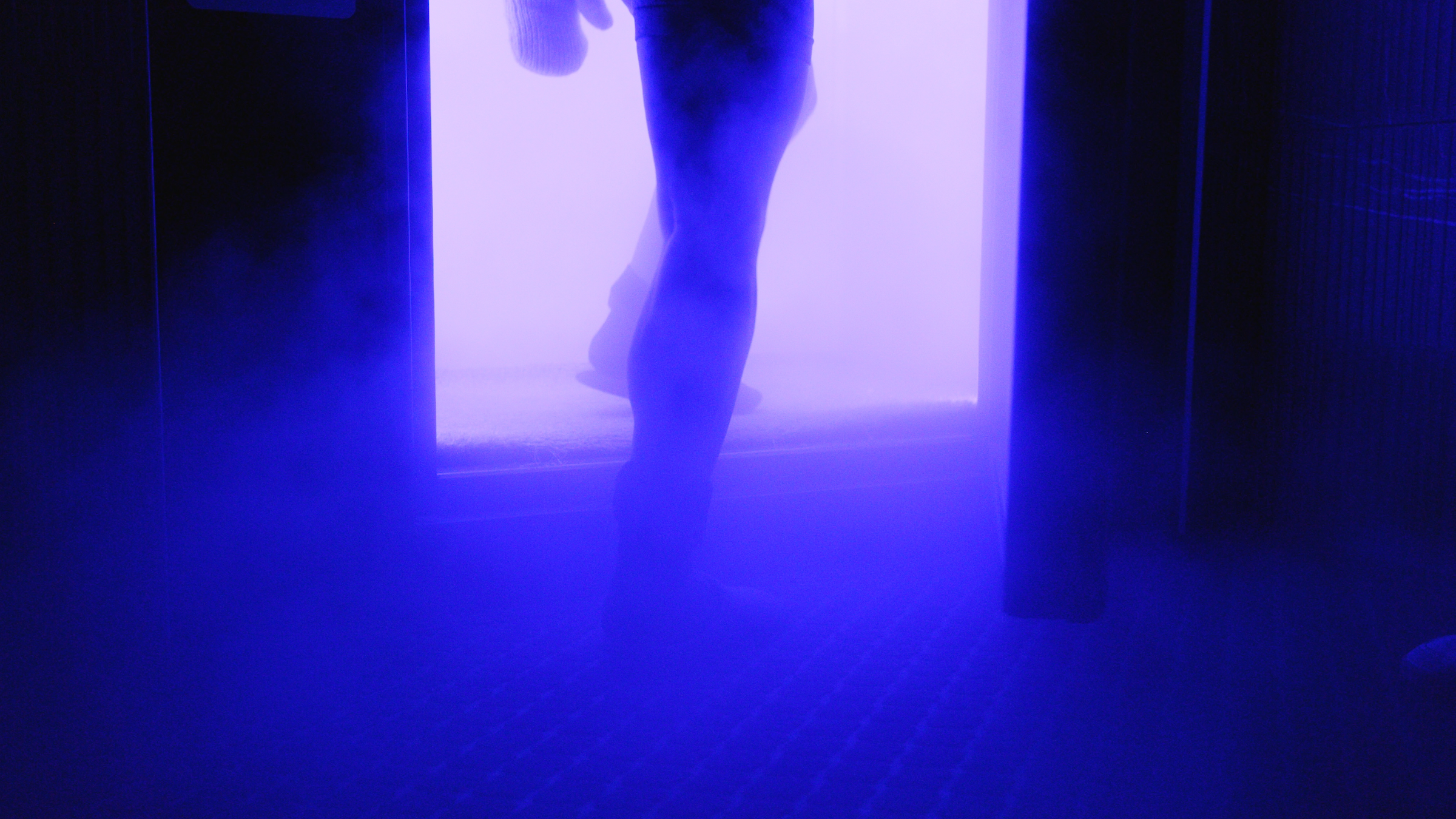 in a blue-lit room, a woman steps into a fogging cryotherapy chamber