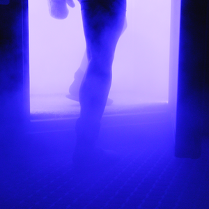 in a blue-lit room, a woman steps into a fogging cryotherapy chamber