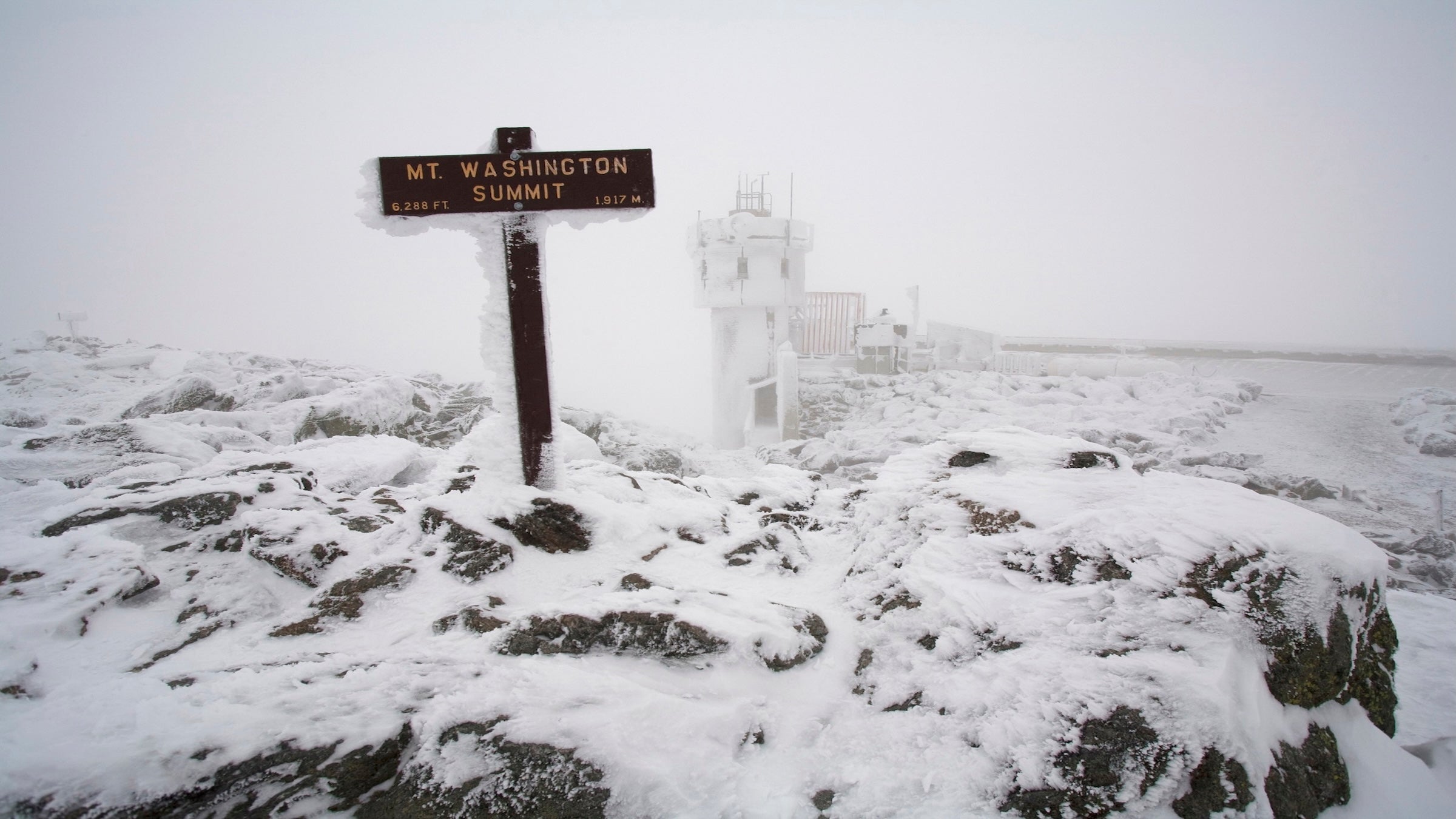 A snow and ice covered sign marks the summit of Mount Washington in the White Mountains of New Hampshire. The Mount Washington Observatory can be seen in the distance.