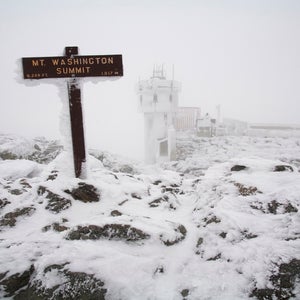 A snow and ice covered sign marks the summit of Mount Washington in the White Mountains of New Hampshire. The Mount Washington Observatory can be seen in the distance.