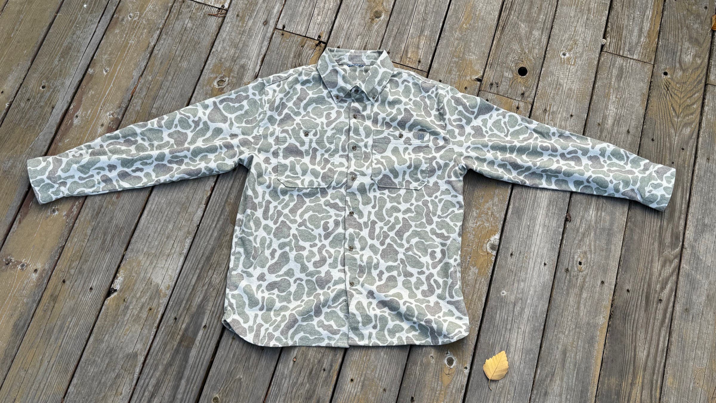 A flat-lay view of the Huk Chesapeake KC Printed Flannel in a grey and white "Army Camo" pattern, showing its dual chest pockets and technical stretch fabric as one of the best flannel shirts for men for fishing or casual wear.