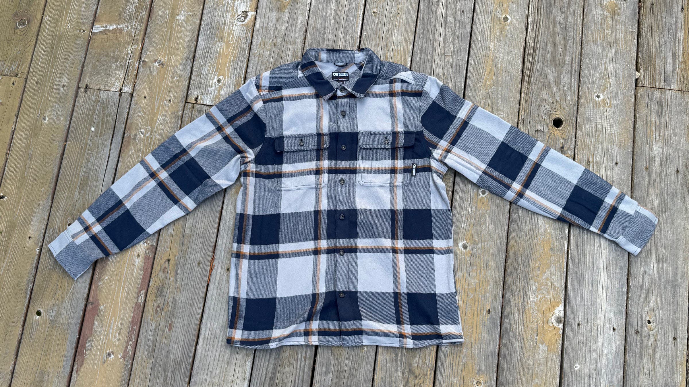 A flat-lay view of the Outdoor Research Feedback Flannel in a blue, white, and tan plaid pattern on a wooden deck, demonstrating why it is one of the best flannel shirts for men seeking technical performance and classic style.