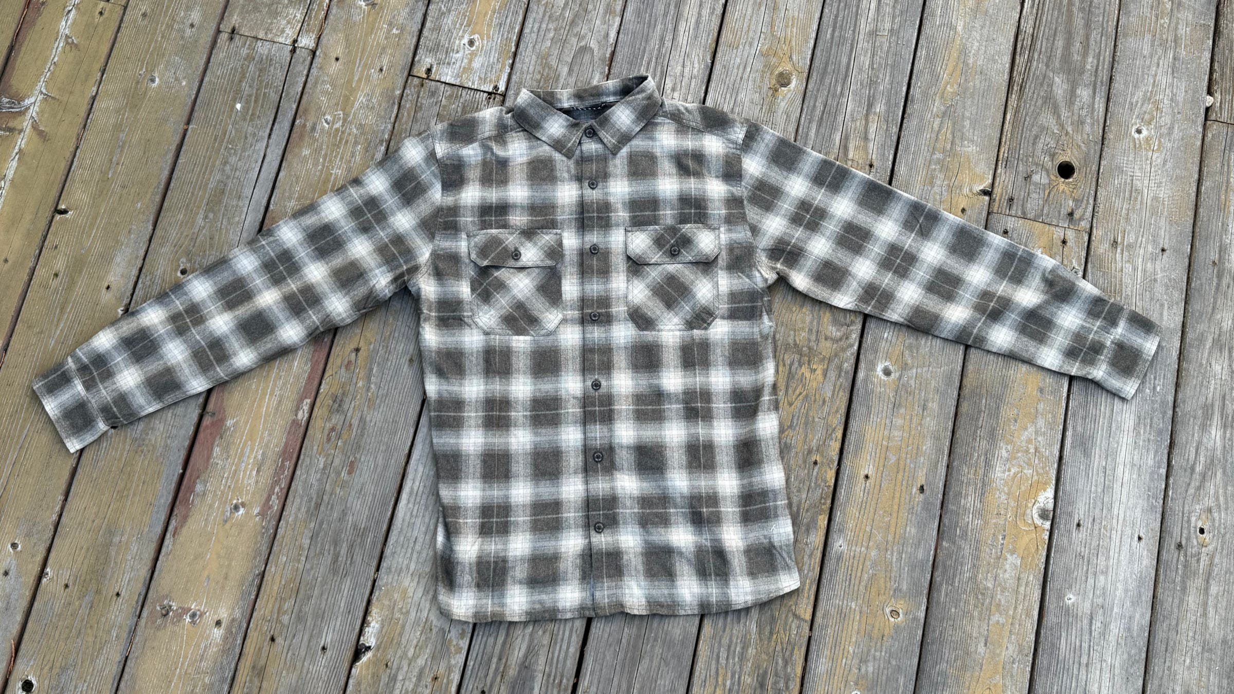 A flat-lay view of the Royal Robbins Men’s Vogelsang L/S on a grey wooden deck, showcasing its grey and white plaid pattern and dual chest pockets as one of the best flannel shirts for men for travel and outdoor versatility.