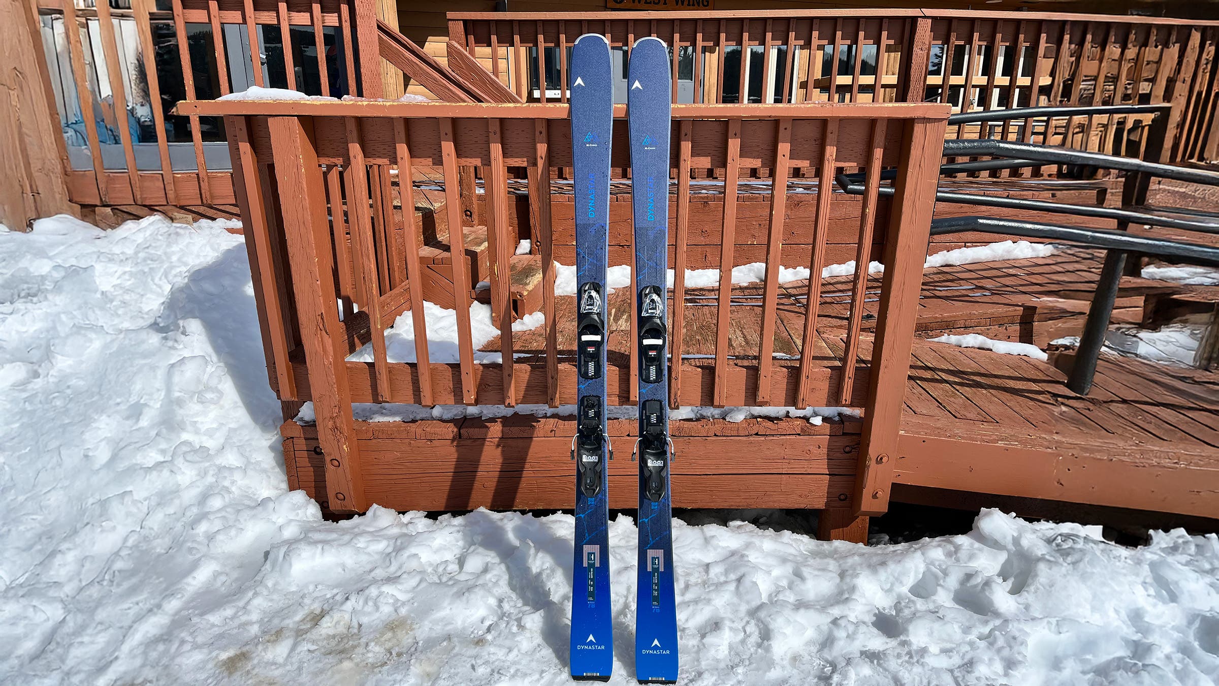 Blue Dynastar M-Cross 78 skis for intermediate skiers propped up on wooden railing