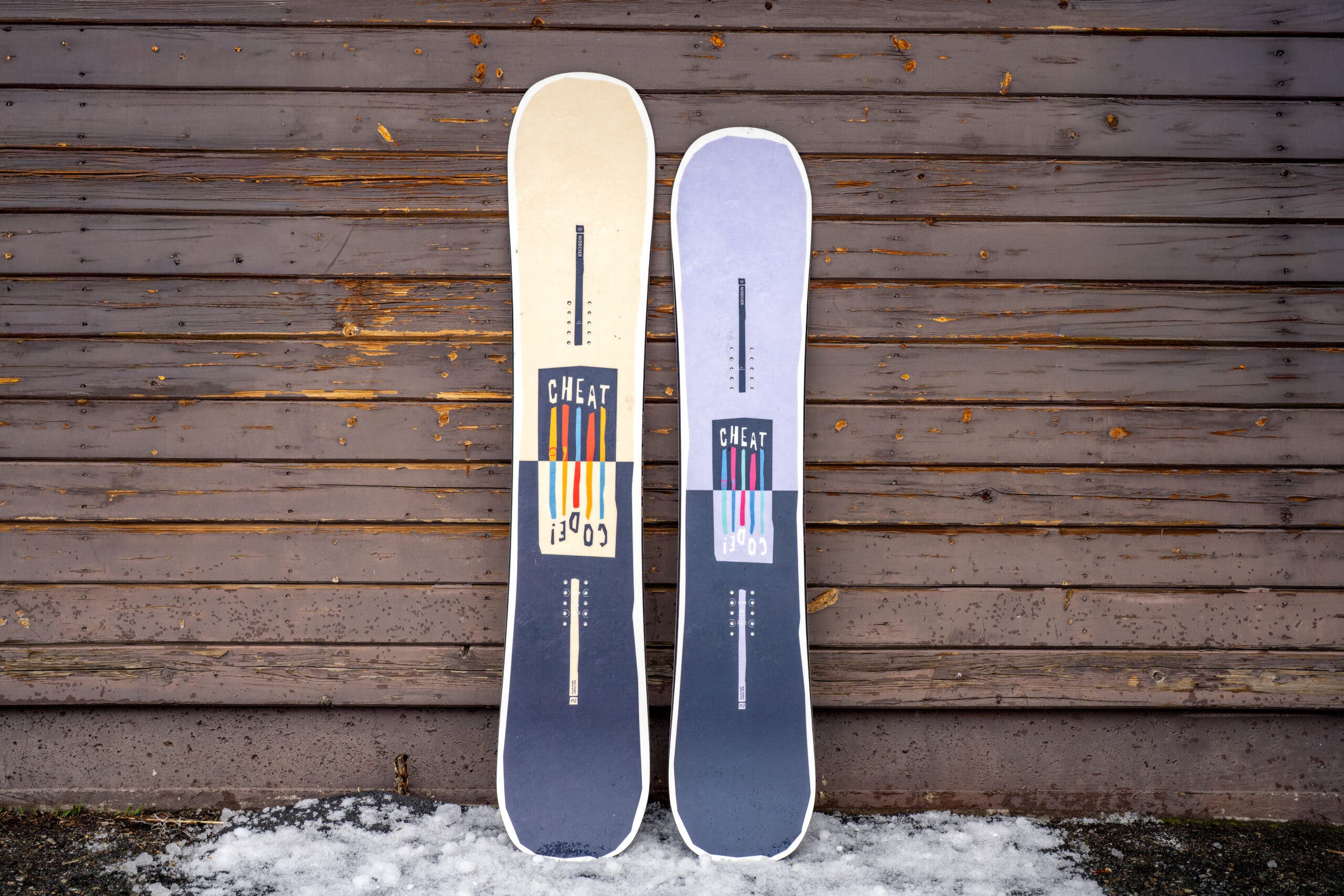 two Nidecker Cheatcode snowboards leaning up against weathered building